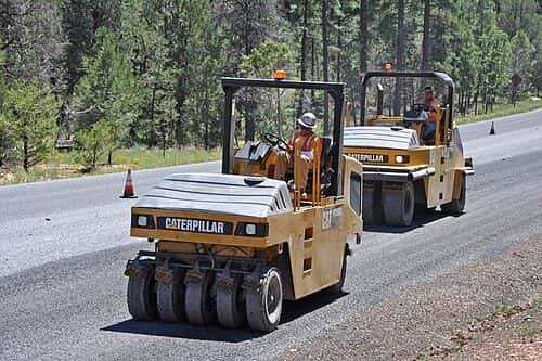 Two rubber-tired rollers compact a fresh chip seal on a two-lane road through pine forest