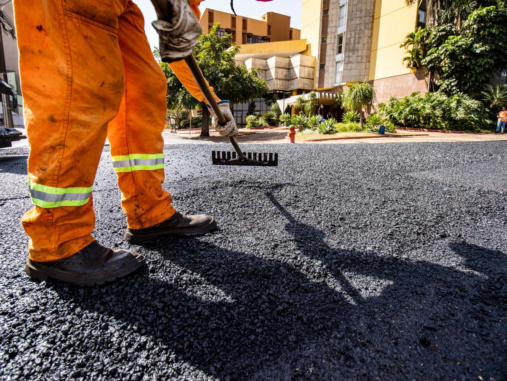 A worker in orange high-visibility pants rakes fresh hot asphalt