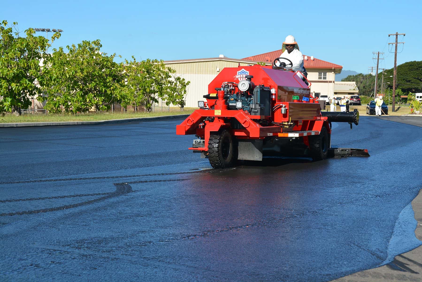 A worker drives a red ride-on sealcoat machine across a freshly sealed parking lot