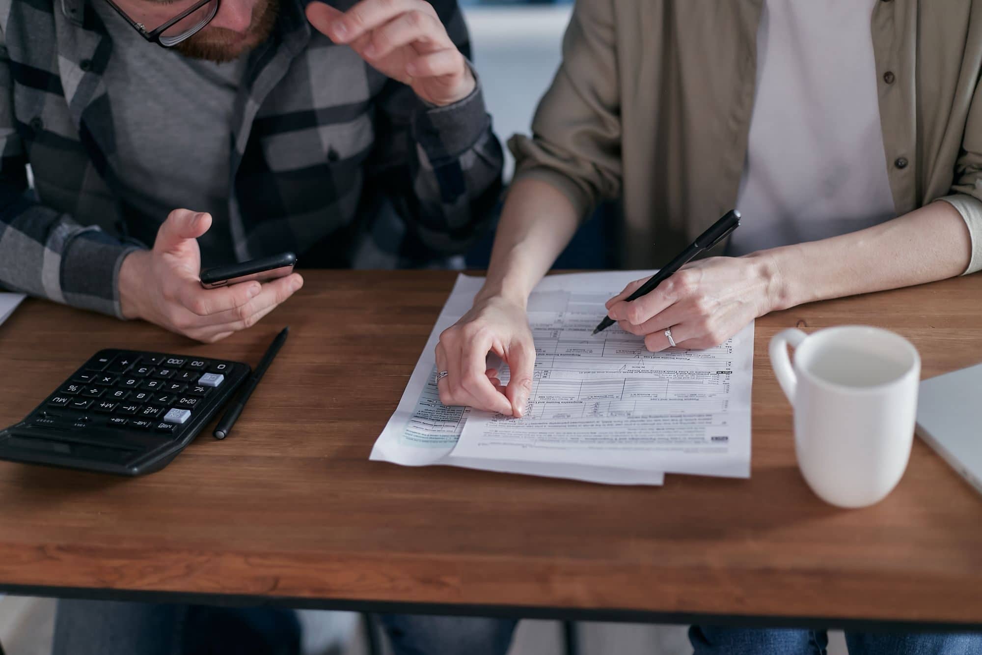 Two people at a table work through tax forms with a calculator and a phone
