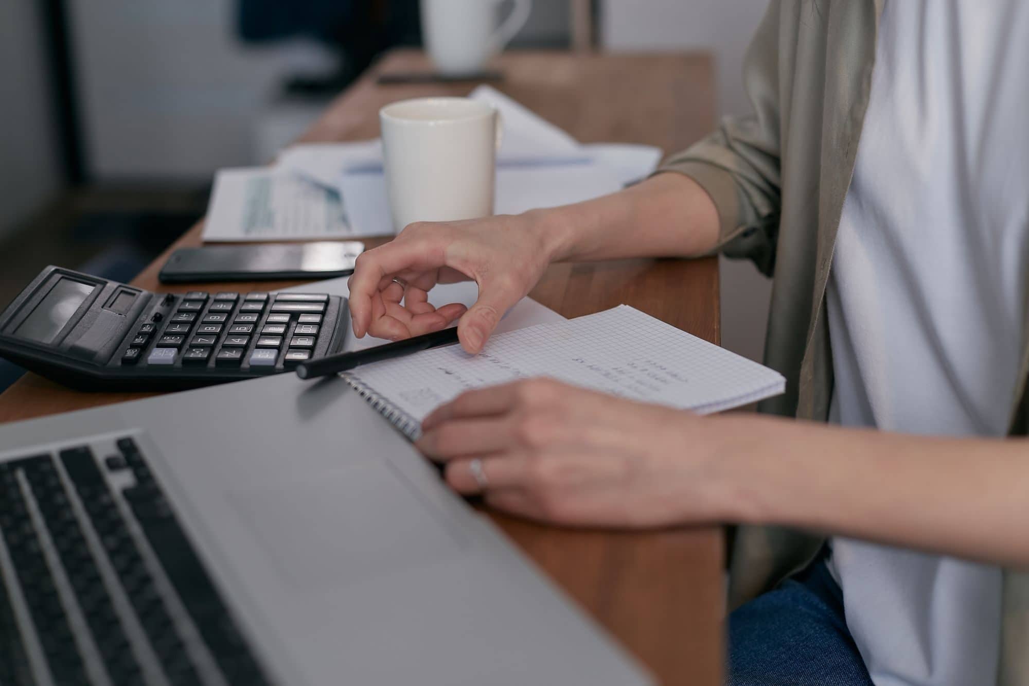 A person writes in a notebook beside a laptop and a calculator