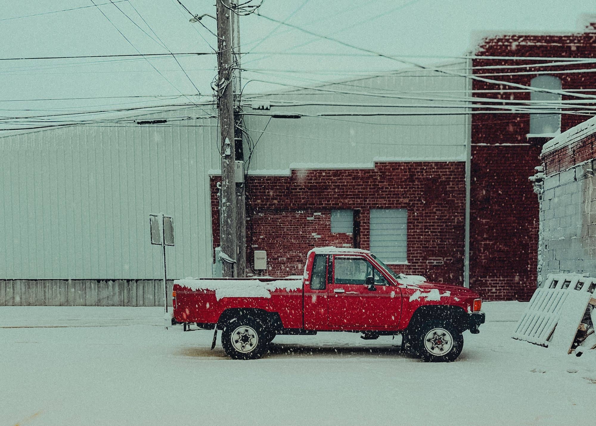 A red pickup truck parked beside a brick building while snow falls