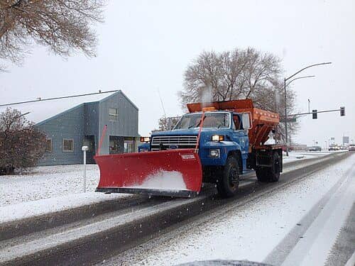 A blue dump truck with a red plow blade and a salt spreader clears a snowy highway