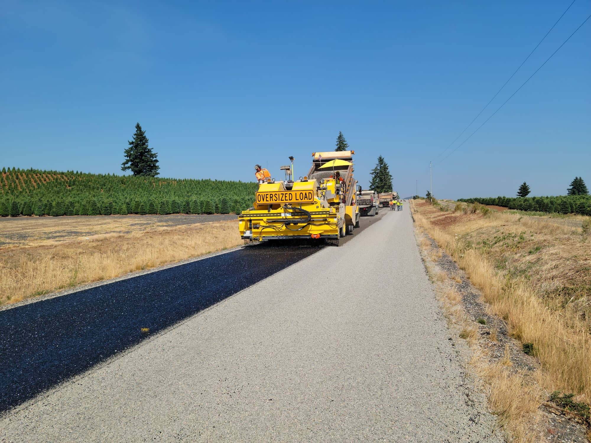 An asphalt paver lays a fresh black strip on a rural path between farm fields