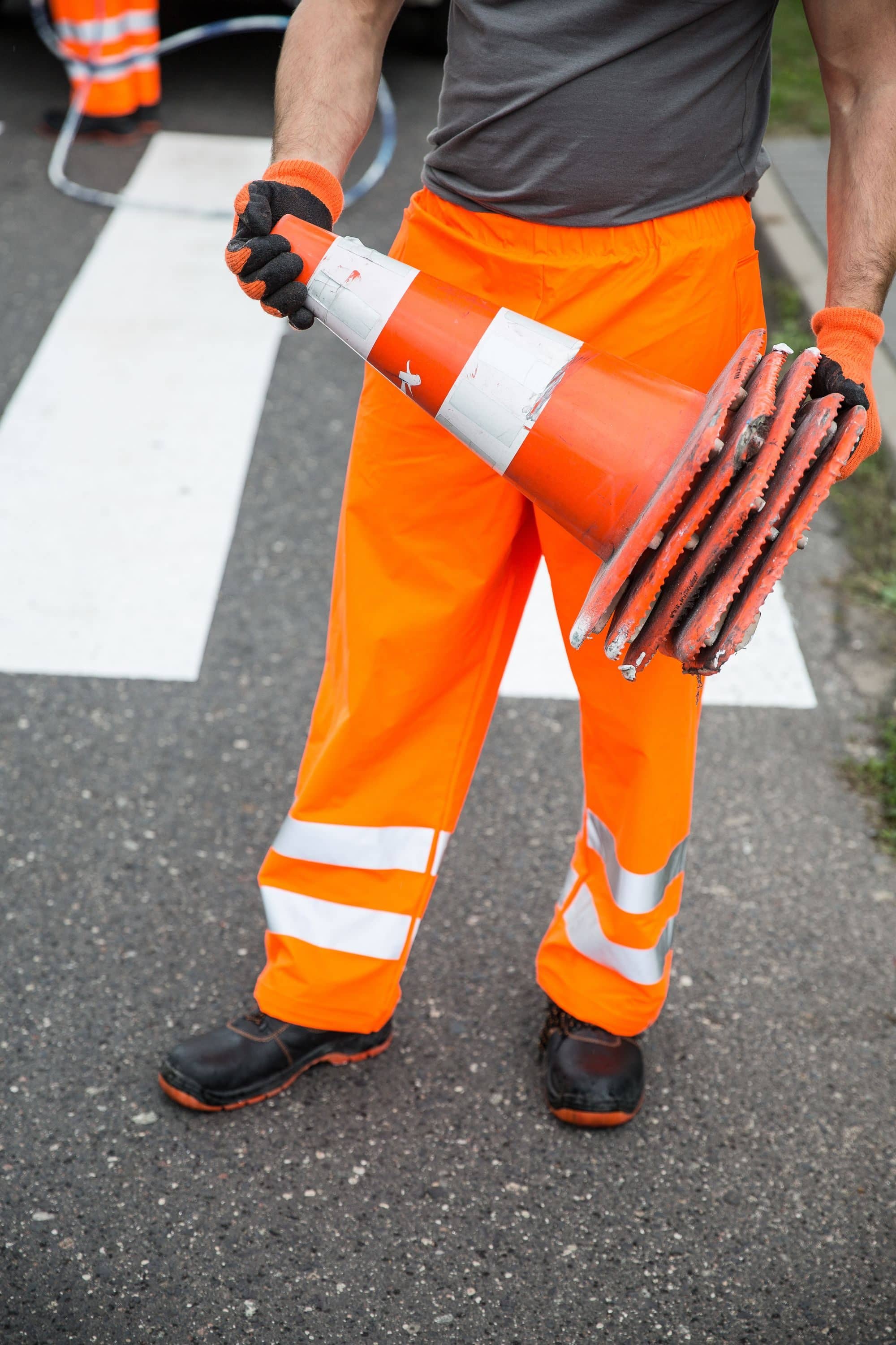 A worker in orange high-visibility pants carries a stack of traffic cones across a crosswalk